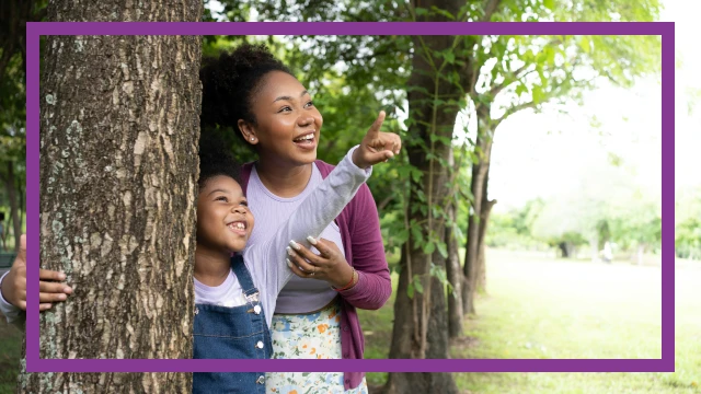 A mother and daughter enjoying a sunny day in the park, surrounded by greenery. The young girl is pointing at something in the distance while she smiles.